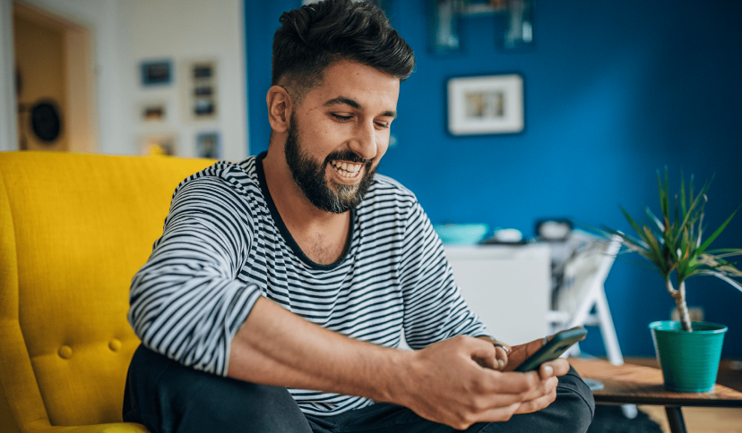 Man in a blue and white striped shirt sitting on a yellow sofa looking at his smartphone at home.