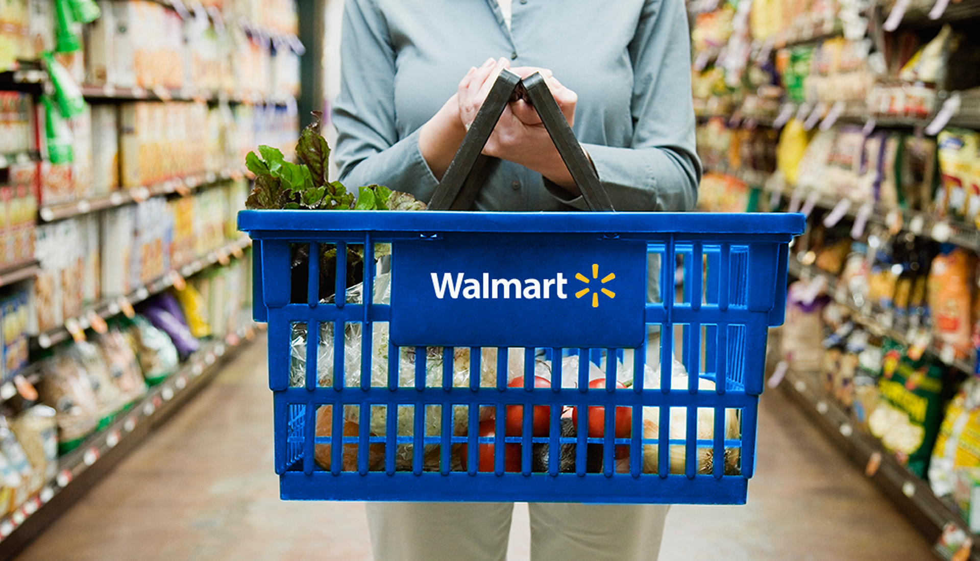 Woman in a light blue cardigan holding a shopping Walmart blue shopping basket in a grocery store aisle.