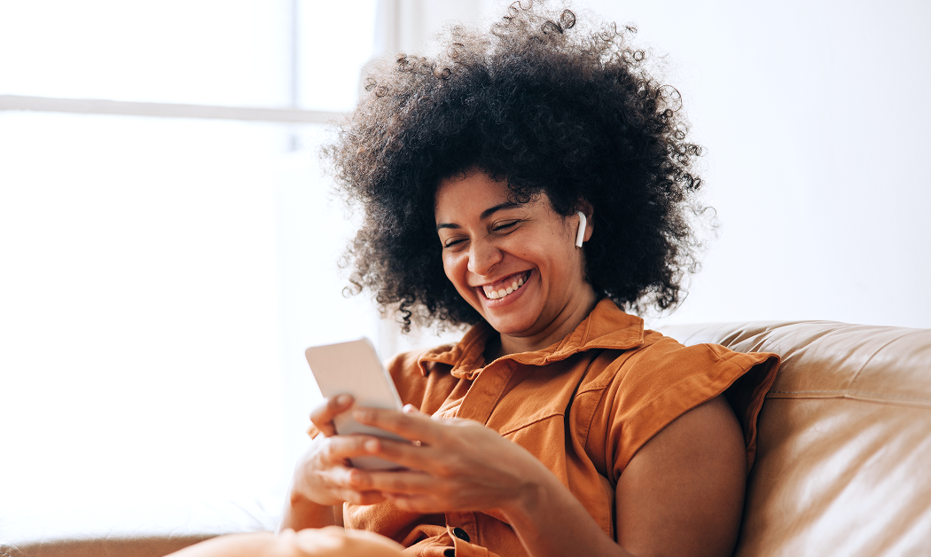 Woman sitting on a brown leather sofa at home with earbuds in looking at her smartphone.