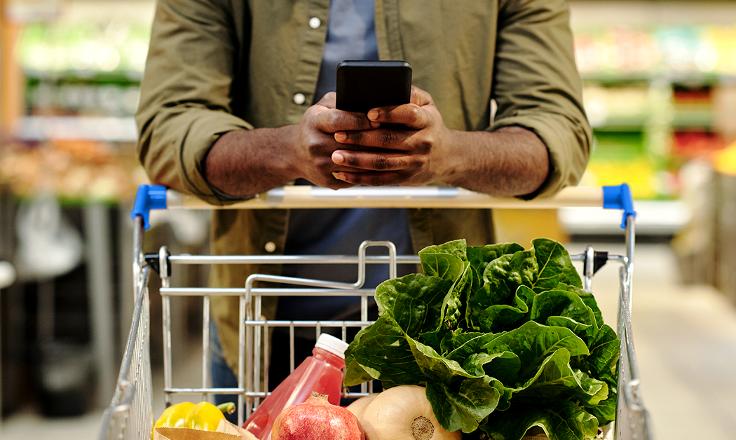 Man looking down at his smartphone as he pushes a Walmart shopping cart filled with groceries.